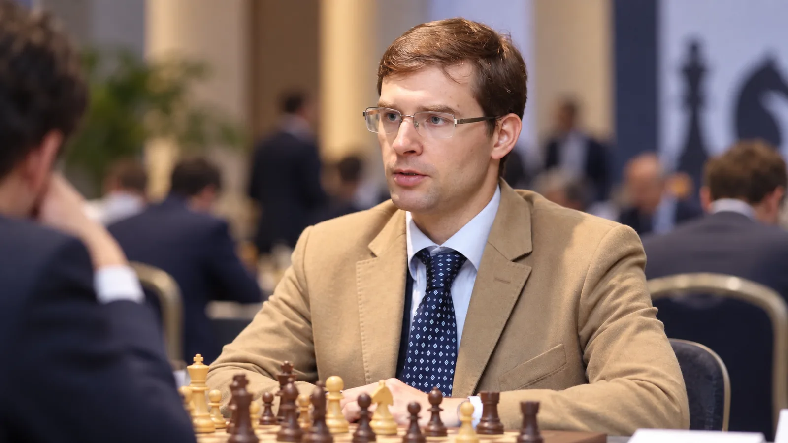 Тимур Турлов в редакционна AI преработка на архивен портрет от април 2023 г. с илюстративен шахматен фон; не е кадър от избора във FIDE. Оригинална снимка: Fassileety / Wikimedia Commons, CC BY-SA 4.0 (https://creativecommons.org/licenses/by-sa/4.0/); източник: https://commons.wikimedia.org/wiki/File:Timur_Turlov.jpg. Преработката се предоставя при CC BY-SA 4.0.