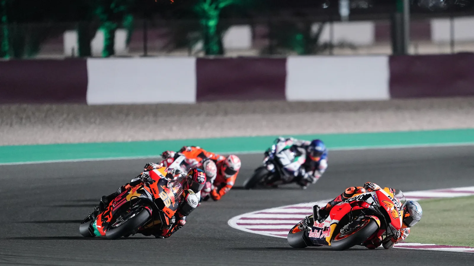 Архив: група пилоти в MotoGP на Гран при на Катар в Лусаил, 28 март 2021 г.; не е кадър от 2026 г. или евакуация. Box Repsol / Wikimedia Commons, CC BY 2.0. Оригинал: https://commons.wikimedia.org/wiki/File:Qatar_GP_2021._Pol_Espargaro_y_Stefan_Bradl_en_carrera_-_51079776027.jpg ; лиценз: https://creativecommons.org/licenses/by/2.0/ ; изрязано и оразмерено до 16:9 без AI редакция.