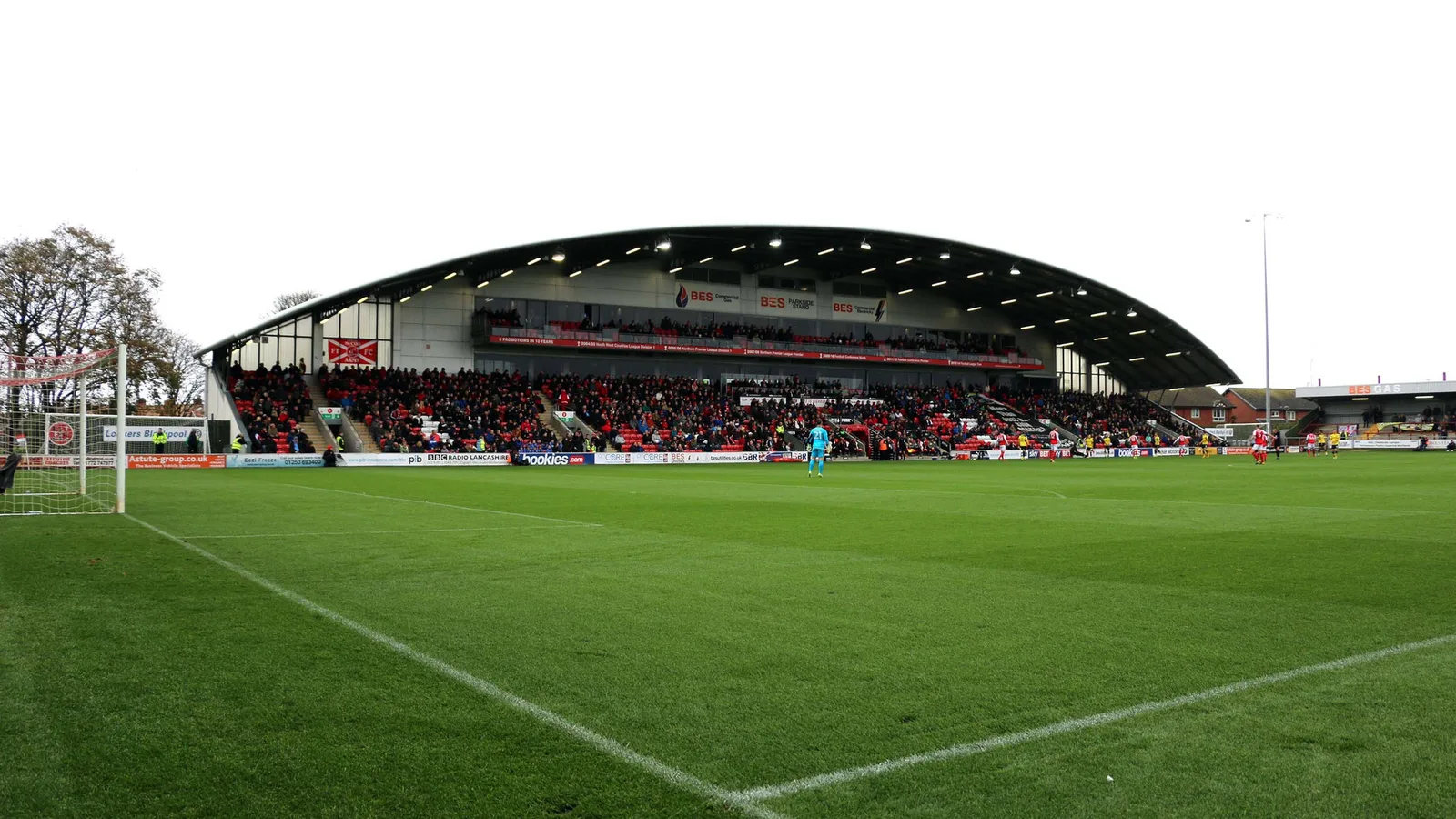 Архив: трибуната Parkside на стадион Highbury във Флийтууд, 28 октомври 2017 г.; не е кадър от Флийтууд – Арсенал през 2026 г. Steve Daniels / Geograph, CC BY-SA 2.0. Изрязано и преоразмерено до 16:9 без AI редакция; адаптацията е под същия лиценз. Оригинал: https://commons.wikimedia.org/wiki/File:The_BES_Parkside_Stand_in_Highbury_Stadium_-_geograph.org.uk_-_5590439.jpg ; лиценз: https://creativecommons.org/licenses/by-sa/2.0/