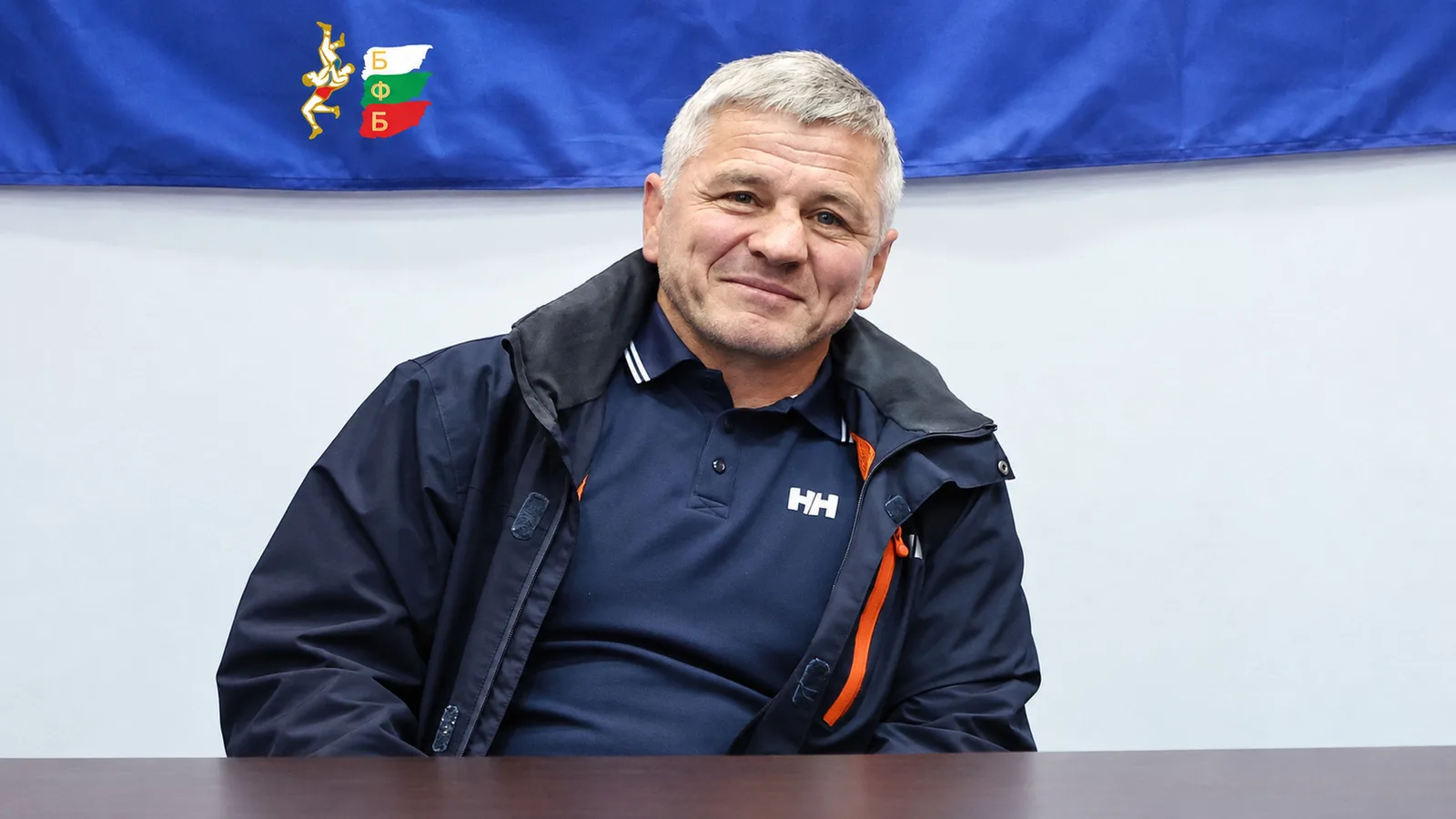Емил Будинов (Бате Емо) — архивен официален портрет на БФ Борба с AI разширение на фона и адаптация 16:9; не е кадър от епизода на „Домът на волята“. Източник: https://bul-wrestling.org/novini/emil-budinov-praznuva-60-godishnina-dnes