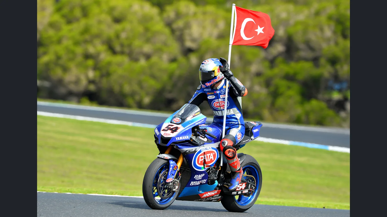 Топрак Разгатлъоглу с турското знаме върху Yamaha по време на WorldSBK на „Филип Айлънд“ през 2020 г. — архивна снимка, не от Мандалика 2026 или MotoGP. Russell Colvin / Wikimedia Commons, CC BY-SA 4.0; оригинал: https://commons.wikimedia.org/wiki/File:Toprak_Razgatl%C4%B1o%C4%9Flu_2020._Image_Russell_Colvin.jpg ; лиценз: https://creativecommons.org/licenses/by-sa/4.0/ ; поставена в 16:9 кадър без изрязване, производното остава под CC BY-SA 4.0.